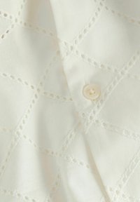 Tessuto bianco con un motivo a diamante e ritagli cuciti. Presenta un piccolo bottone rotondo di colore crema sul bordo.