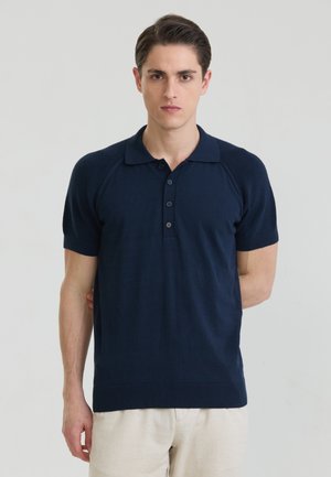 Fiatal férfi rövid sötét hajjal, sötétkék rövid ujjú galléros pólót és világos nadrágot viselve, egyszínű háttér előtt áll.