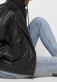 Chaqueta de cuero negro con un acabado texturizado, que cuenta con cierre de cremallera y detalles de botones a presión, combinada con vaqueros azul claro.