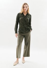 Camicia button-up verde scuro con stampa leopardata, abbinata a pantaloni larghi color oliva chiaro e ballerine beige. Tessuto liscio e leggero.