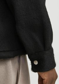 Veste en laine noire avec un tissu texturé, dotée d'un poignet arrondi et d'un détail de bouton-pression métallique au niveau de la manche.