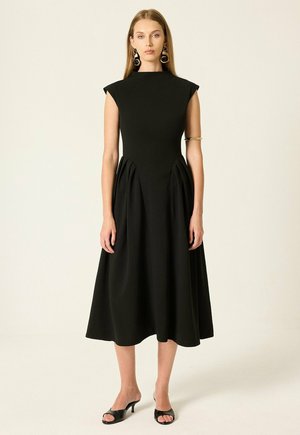 Femme portant une robe midi noire sans manches avec épaules rembourrées, jupe plissée, sandales noires à talons, grandes boucles d'oreilles rondes et un bracelet manchette en or.