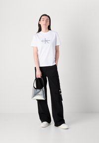 Camiseta blanca de algodón con el logotipo de "Calvin Klein", combinada con pantalones cargo negros con bolsillos y un bolso plateado reflectante. Zapatillas blancas.