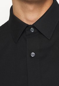 Svart skjorta med krage och knäppning. Har en texturerad tygkvalitet och två mörka knappar med fyra h ål vardera, släta kragekanter.