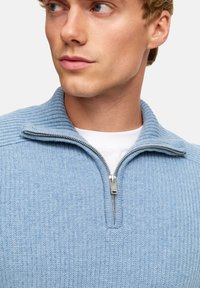 Jasnoniebieski sweter zapinany na zamek z prążkowanym wzorem, stójką i srebrnym zamkiem. Nosi się go na białej koszulce z okrągłym dekoltem.