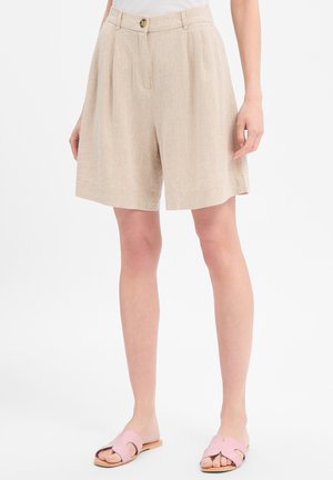 Femme portant un short plissé beige taille haute avec bouton et fermeture éclair, associé à des sandales slides rose clair, debout sur fond blanc.
