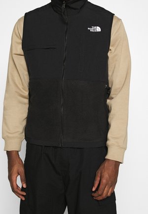 Mężczyzna ubrany w czarną kamizelkę polarową na zamek z logo The North Face, na beżowej koszuli z długim rękawem i czarnych spodniach, stojący z ramionami luźno opuszczonymi.