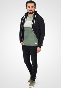 Sort zip-up hoodie med hvide snore, lagdelt over en grøn T-shirt med grå stribemønster. Sorte slim-fit jeans og grå sneakers.