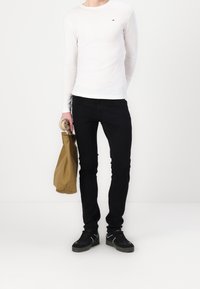 Persona con una camiseta de manga larga blanca, pantalones negros y zapatillas negras, sosteniendo un paraguas enrollado y una bolsa marrón frente a un fondo liso.