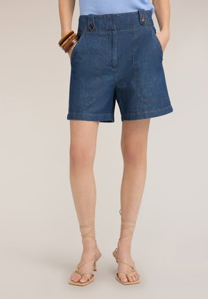Kvinde iført mørkeblå cowboyshorts med højtaljede og forlommer, kombineret med beige strop-sandaler med hæl og træ-armbånd.