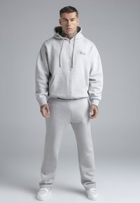Grijze hoodie met rits en trekkoorden, gecombineerd met bijpassende joggingbroek. Zachte stof, losse pasvorm, met geborduurde details op de borst en het been.