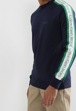 Mężczyzna noszący granatową bluzę z zielono-białymi paskowanymi rękawami z napisem „Calvin Klein Jeans”, w połączeniu z beżowymi spodniami.