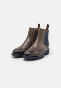 Par de botas de tornozelo em couro castanho com detalhes de brogue, painéis elásticos laterais escuros, alças para puxar e solas texturizadas em azul-marinho, sobre fundo branco.