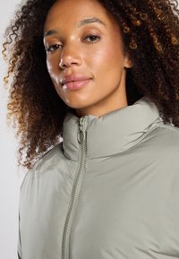 Chaqueta acolchada verde claro con un alto cuello y textura suave. Cuenta con un cierre frontal y un detalle elástico en el cuello.