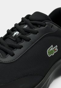 Κοντινό πλάνο μαύρου αθλητικού παπουτσιού με υφή από πλεκτό δίχτυ, μαύρα κορδόνια και πράσινο κεντημένο λογότυπο κροκοδείλου στη πλευρά.