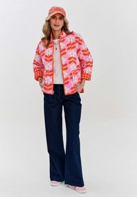 Prešívaná bunda s ružovým kvetinovým vzorom, oranžovými akcentmi a zipsom, v kombinácii s bielou košeľou a širokými tmavými denimovými nohavicami.