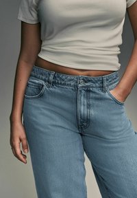 Svetlo modri denim hlače s sproščenim krojem, zasnovo s petimi žepi in gumbnim zapiranjem, v kombinaciji z oprijetim belim topsom z rebrasto strukturo.