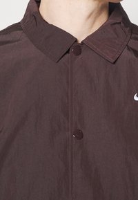 Burgundská lehká bunda s knoflíkovým zapínáním, špičatým límcem a malým bílým logem na levém prsu. Hladká texturovaná látka.