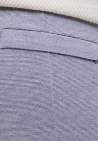 Helles blaufarbenes Gewebe mit einer strukturierten Webart. Verfügt über eine Gesäßtasche und einen glatten Bund. Der obere Rand ist teilweise von einem weißen Kleidungsstück bedeckt.