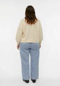 Femme avec des cheveux bruns ondulés portant un pull crème à manches texturées, un jean large bleu clair et des chaussures noires, se tenant de dos.