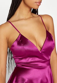Σατέν φόρεμα σε σκούρο ματζέντα με V-λαιμόκοψη, λεπτές ρυθμιζόμενες straps και εφαρμοστό κορσέ, διαθέτοντας μία λεία, γυαλιστερή υφή.