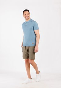 Polo a maniche corte blu chiaro con tre bottoni, abbinato a pantaloni cargo verde oliva, indossato con sneakers bianche, su uno sfondo semplice.