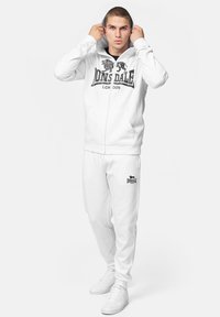 White zip-up hoodie with black logo, kangaroo pockets, matching sweatpants, and white sneakers. Soft fabric with smooth texture.