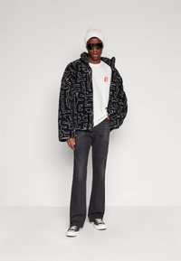 Chaqueta de forro polar con patrón negro y gris, camiseta blanca, jeans oscuros y zapatillas negras. Los accesorios incluyen un gorro de punto blanco y gafas de sol.