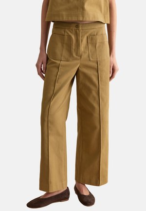 Donna con pantaloni beige a gamba larga con tasche applicate frontali e scarpe marroni basse, in piedi davanti a uno sfondo semplice.
