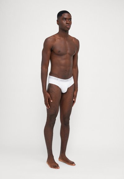 Culotte blanche avec une ceinture élastique douce arborant un logo de marque gris. Le modèle se tient pieds nus, devant un fond neutre.
