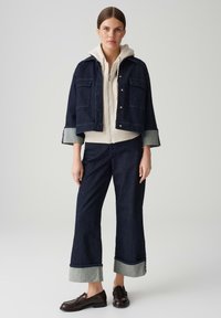 Giacca di jeans scura con tasche a pattina, indossata sopra un felpa grigia chiara. Abbinata a pantaloni di jeans larghi scuri con orli a contrasto. Scarpe marroni.