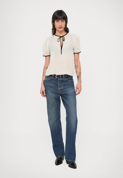 Vit texturerad topp med puffiga ärmar och svart kant, med ett band som knyts i halsringningen. Bärs med blå jeans med raka ben och svarta stövlar.