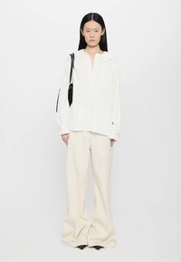 Cămașă albă cu mânecă lungă și guler, asociată cu pantaloni largi crem. Modelul poartă o geantă de mână neagră. Fundal simplu.