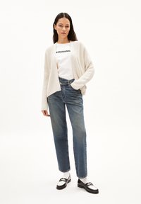 Lichtbeige gebreide cardigan over een wit T-shirt met zwarte tekst. Blauwe high-waisted jeans met een relaxte pasvorm, gecombineerd met zwart-witte loafers.