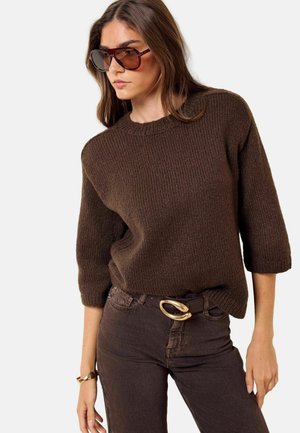 Frau trägt braunen Strickpullover, dunkelbraune Hose mit Gürtel mit goldener Schnalle, große Sonnenbrille und goldenes Armband, steht vor einfarbigem Hintergrund.