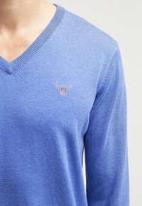 Blaue V-Ausschnitt-Strickjacke aus weichem Material. Mit dem Markenlogo "GANT" und einem kleinen Wappenemblem auf der linken Brust.