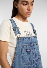 Los overoles de mezclilla azul cuentan con tirantes ajustables, un bolsillo frontal con un parche de logo y un corte relajado sobre una camiseta de punto crema.