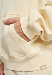 Cremefarvet hoodie med en foran lomme, ribbede manchetter og en lille mærkelap med teksten "DEF" nær kanten. Blød tekstur, afslappet design.
