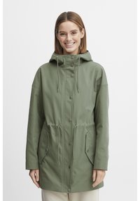 Veste imperméable vert olive avec capuche à cordon de serrage, boutons-pression, taille élastique et deux poches latérales. Tissu lisse et léger.