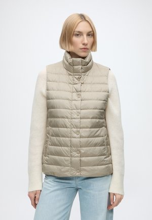 Frau mit kurzen blonden Haaren, die eine beige gesteppte ärmellose Weste über einem weißen Langarmpullover und hellblauen Jeans vor einem einfarbigen Hintergrund trägt.