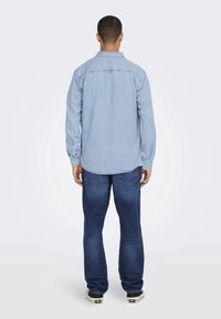 Camicia in denim azzurro chiaro con maniche lunghe e colletto a botones, abbinata a jeans blu scuro e sneakers nere, vista da dietro.