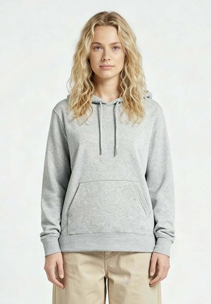 Blonde vrouw met golvend haar, gekleed in een lichtgrijze hoodie en beige broek, staand tegen een effen lichte achtergrond.