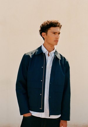 Noormees lokkis juustega, seljas tumesinine jakk valge nööpidega särgi kohal, seisab tavalise beeži seina ees ja vaatab kõrvale.