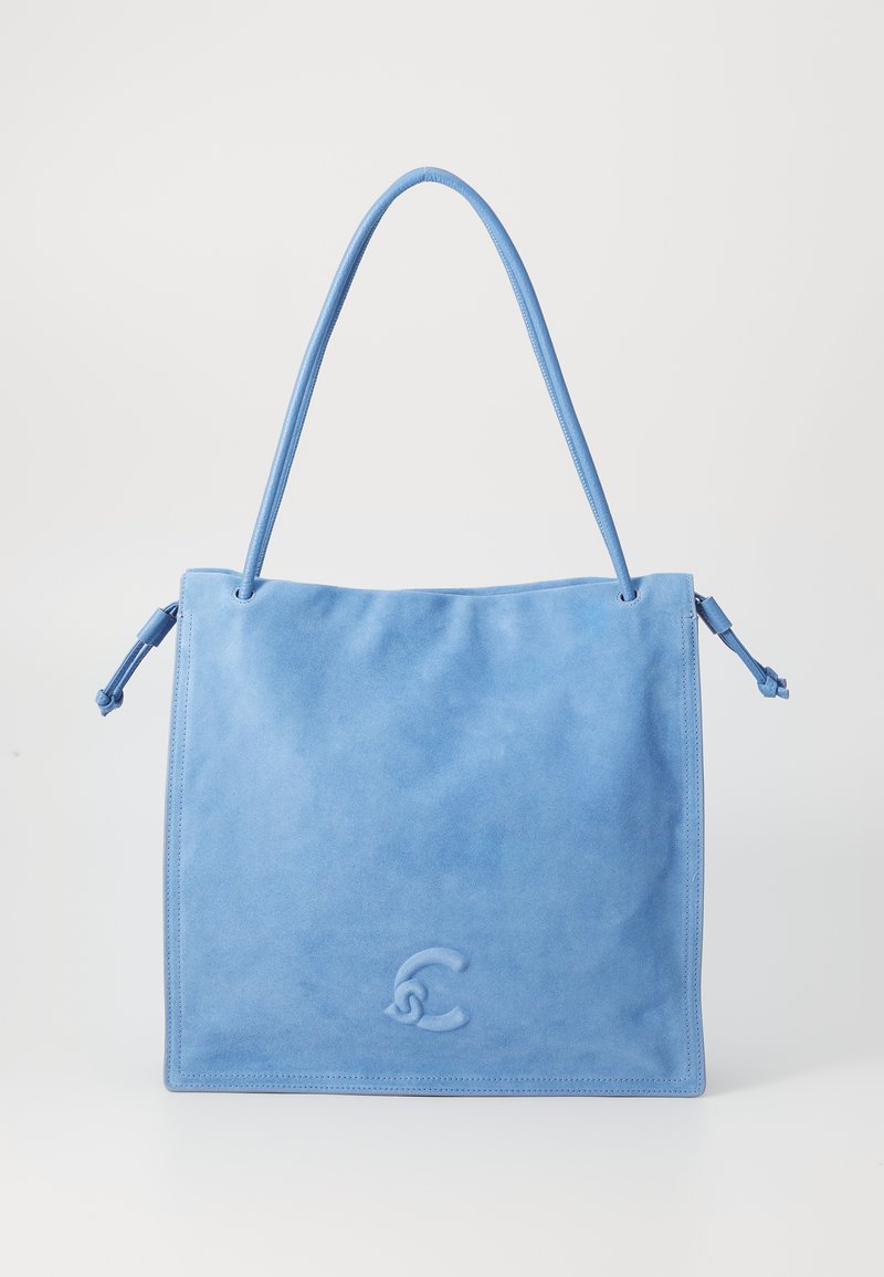 Sac fourre-tout bleu en suède avec une texture lisse, de forme rectangulaire, deux longues poignées et un logo légèrement en relief à l'avant.