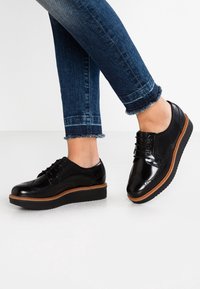Chaussures à lacets en cuir noir avec un design perforé, une semelle plate basse et un accent brun clair autour de la semelle. Portées avec un jean bleu.
