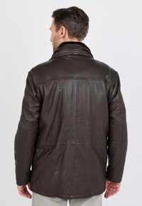 Man met zijn rug naar de kijker, gekleed in een donkerbruin leren jack met zichtbare stiksels en een kraag van bont, staand tegen een effen achtergrond.