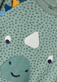 Camicia di tessuto verde chiaro con un design di faccia di dinosauro; presenta macchie testurizzate, occhi ricamati e accenti triangolari colorati sul colletto.