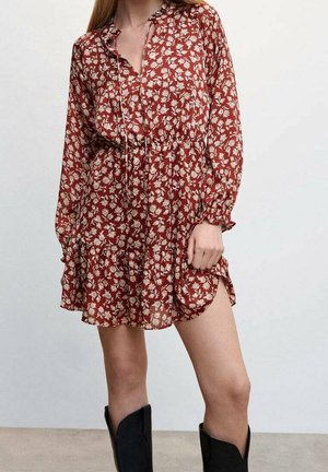 Femme portant une robe longue à manches longues fleurie rouge rouille avec un ourlet à volants et des bottes noires montantes jusqu’aux genoux, debout sur un fond uni.