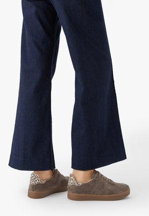 Person iført mørkeblå vide jeans og brune ruskindssneakers med leopardmønstret hæl, stående på en hvid baggrund.