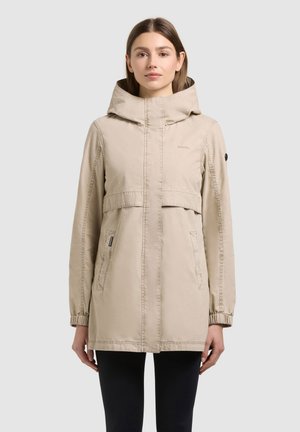 Kobieta w beżowej kurtce z kapturem, zapinanej na zamek błyskawiczny, z kieszeniami na klapki, stojąca na jednolitym jasnoszarym tle.